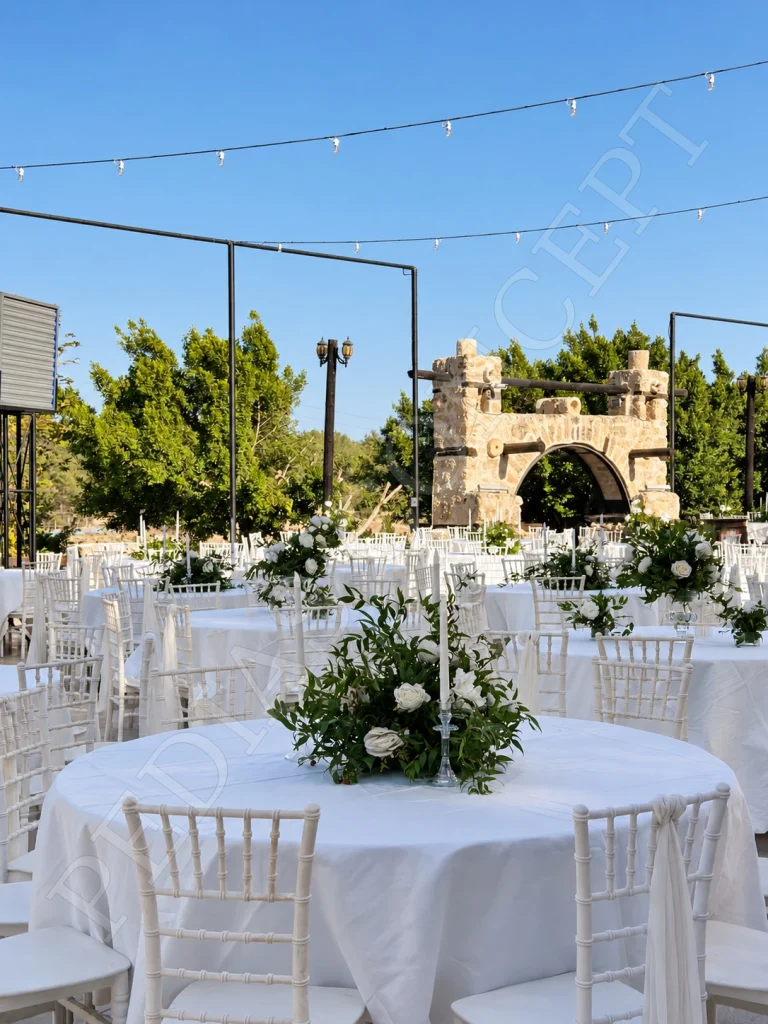 Pedias Concept Olympia Açık Alan’da beyaz çiçeklerle süslü yuvarlak masalar ve arkadaki taş kemer