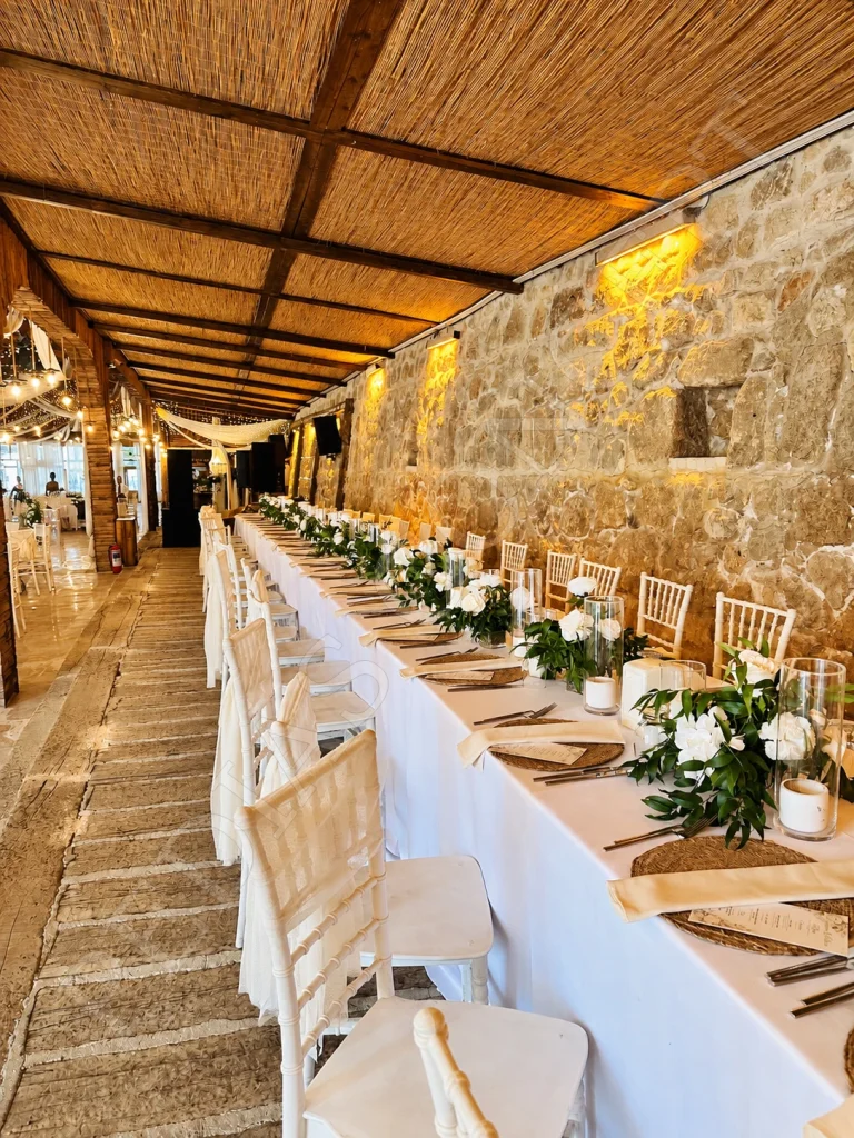 Pedias Concept Hera Kapalı Alan’da taş duvar boyunca çiçeklerle hazırlanan uzun davet masası